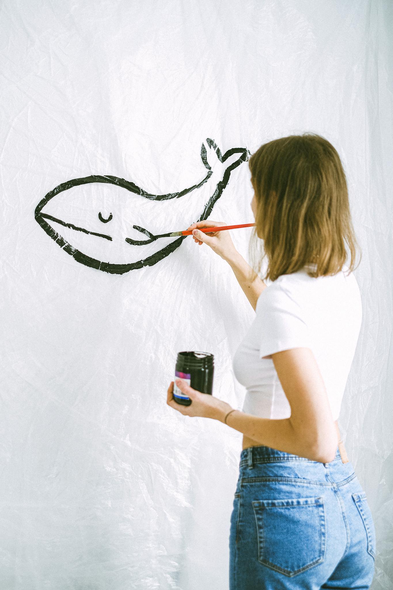 A woman painting a black whale silhouette on a white canvas with brush and ink.