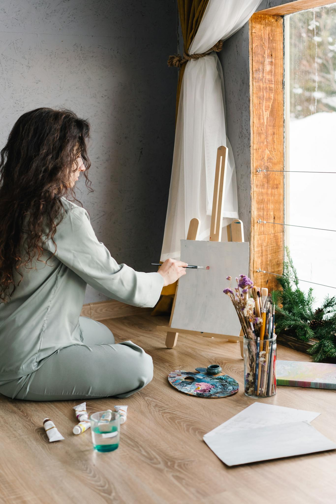 Woman in sleepwear painting on canvas by window with winter view.